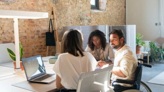 man in a wheelchair talking to two women at desk