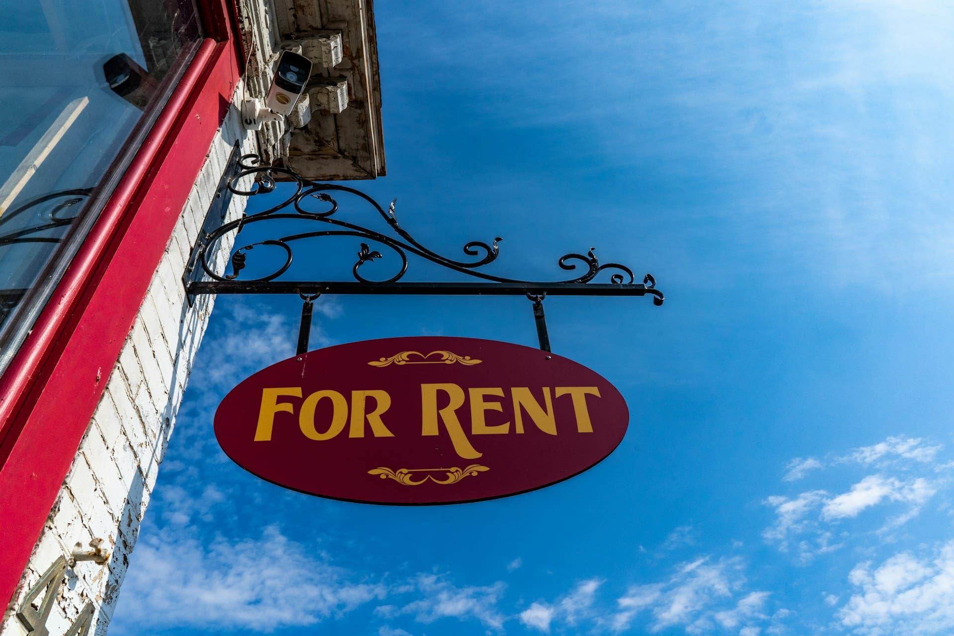 Red and yellow 'For Rent' sign hanging from a building with a clear blue sky behind it.