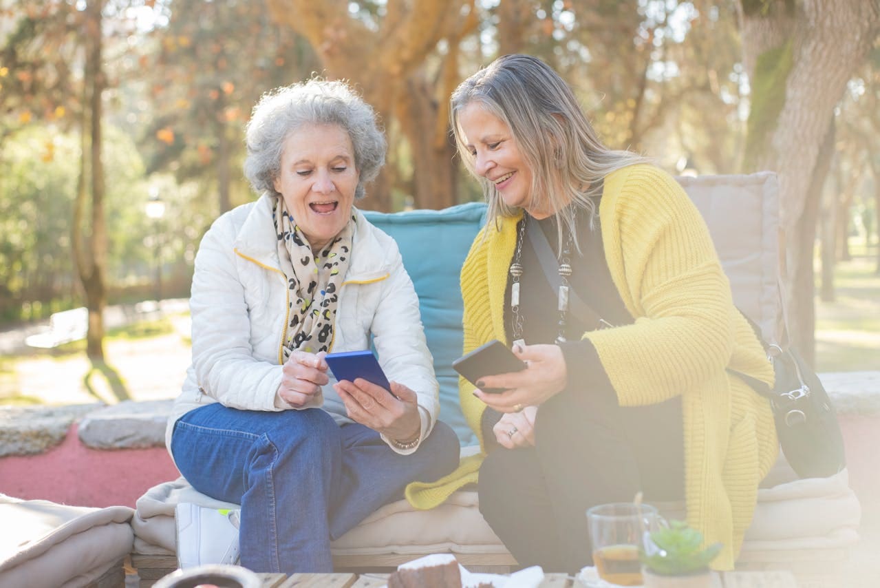 Elderly mother and adult daughter sitting on a bench looking at their phones and laughing